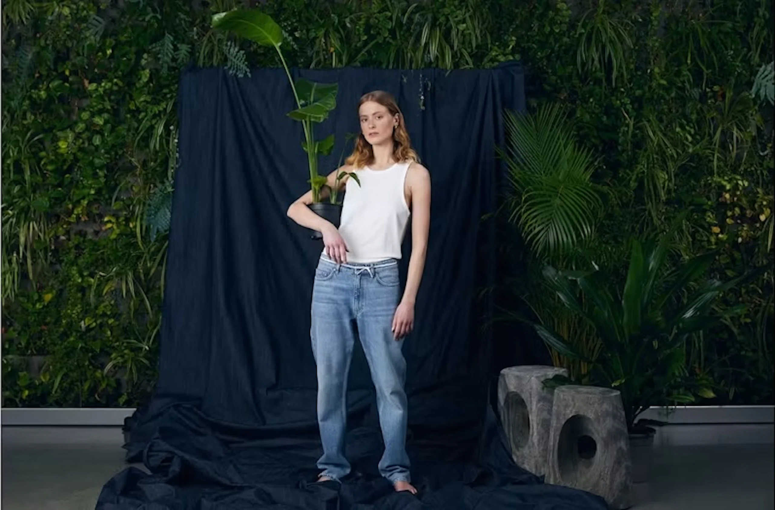 Ung kvinna i vitt linne och lösa jeans som håller en krukväxt med höga gröna blad, stående mot en mörk draperad bakgrund med växtlighet.