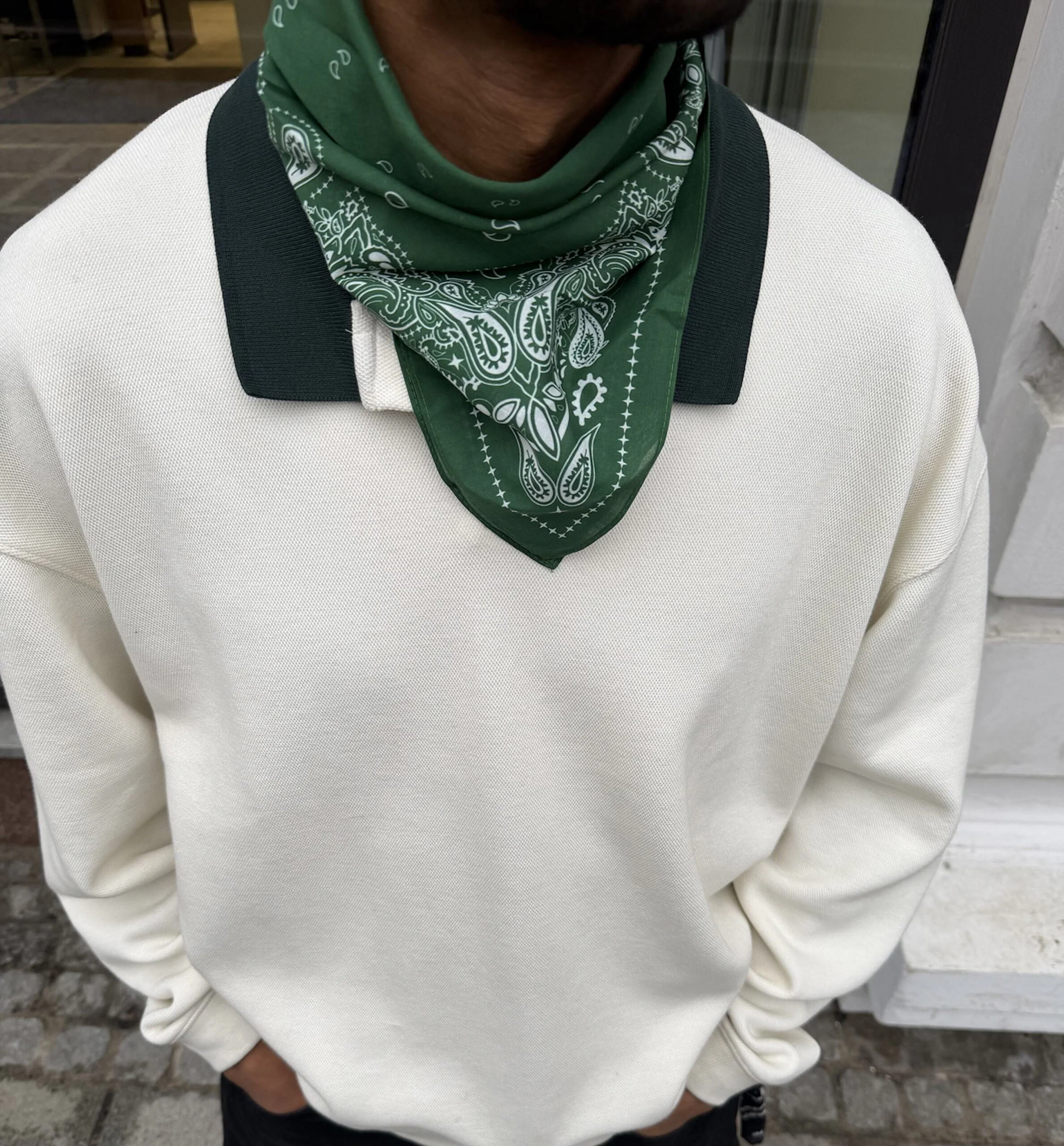 Person bär en vit sweatshirt med mörk krage och en grön bandana med paisleymönster runt halsen.