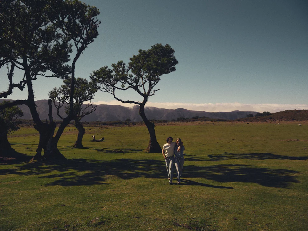 Couple walking arm-in-arm across a sunlit grassy field with twisted trees in foreground and low mountains under a clear sky in the background.