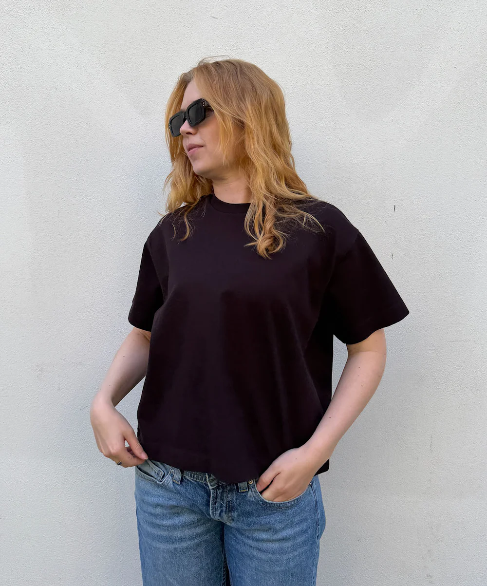 Kvinne med langt rødt hår og solbriller iført mørk oversized t-skjorte og lyse jeans, foran en hvit vegg.