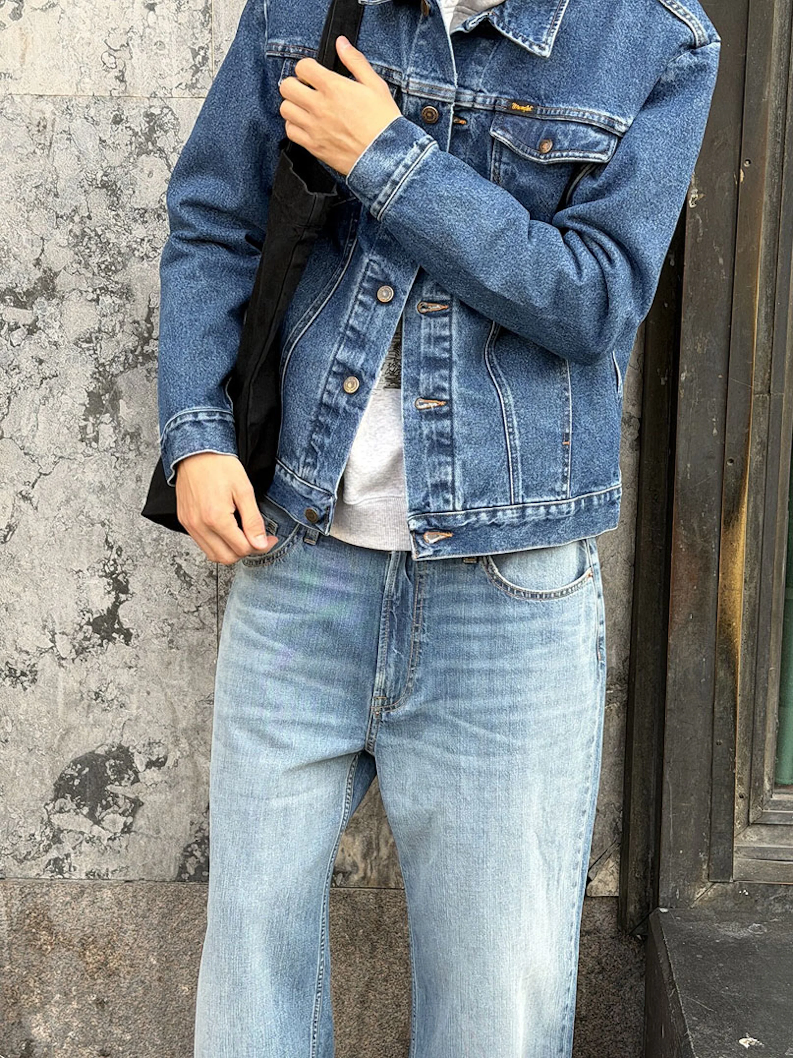 Person i blå jeansjacka och ljusblå jeans, håller en svart väska, står mot en grå stenvägg.